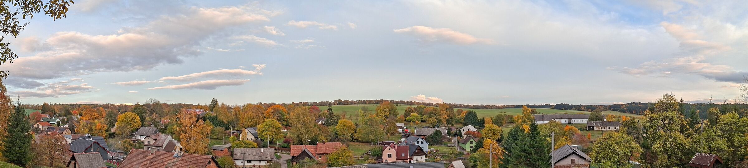 Skuhrov na podzim: střechy domů, barevné stromy, za vsí pole a&nbsp;les.